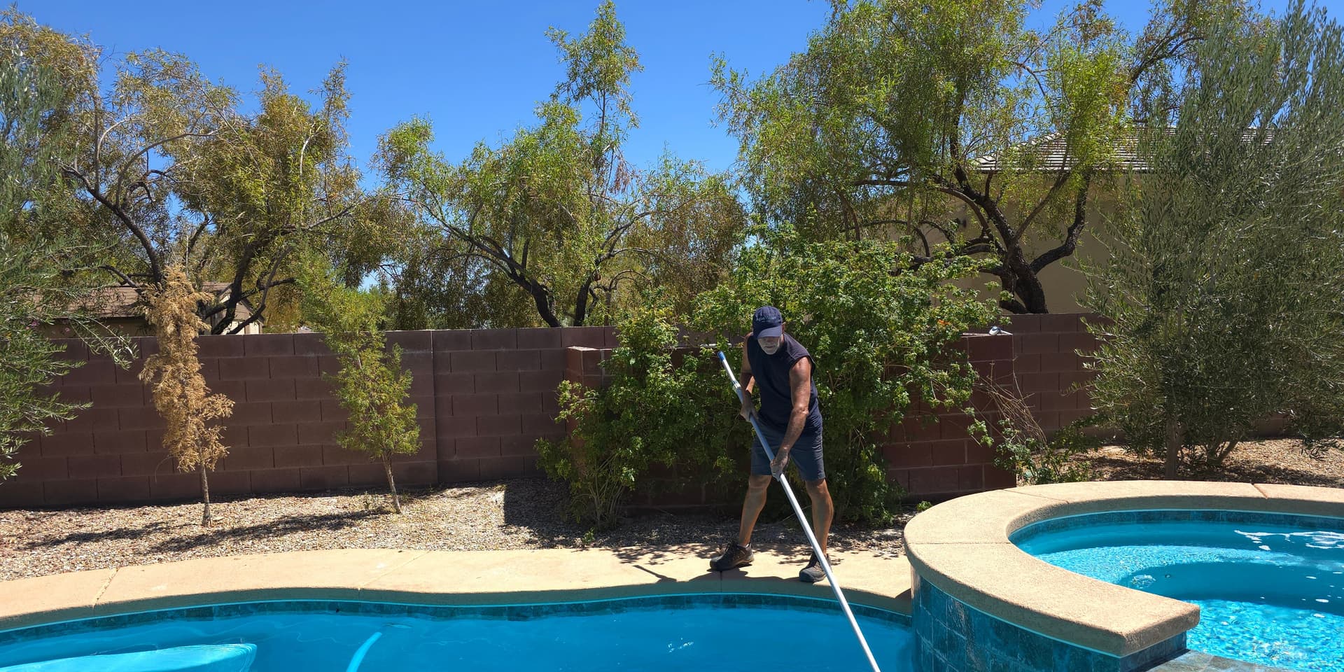 Michael Brown, AquaVision Pool Care Certified Pool Operator, skimming a residential pool and spa with desert trees in North Las Vegas