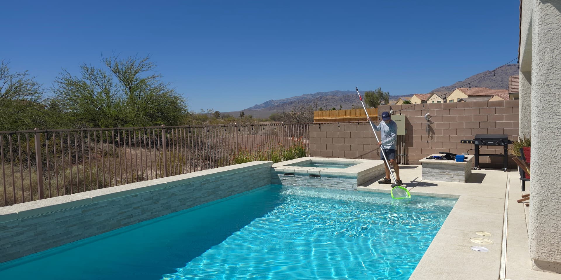 Michael Brown, AquaVision Pool Care Certified Pool Operator, skimming a Northwest Las Vegas pool and spa with desert landscape and mountain backdrop