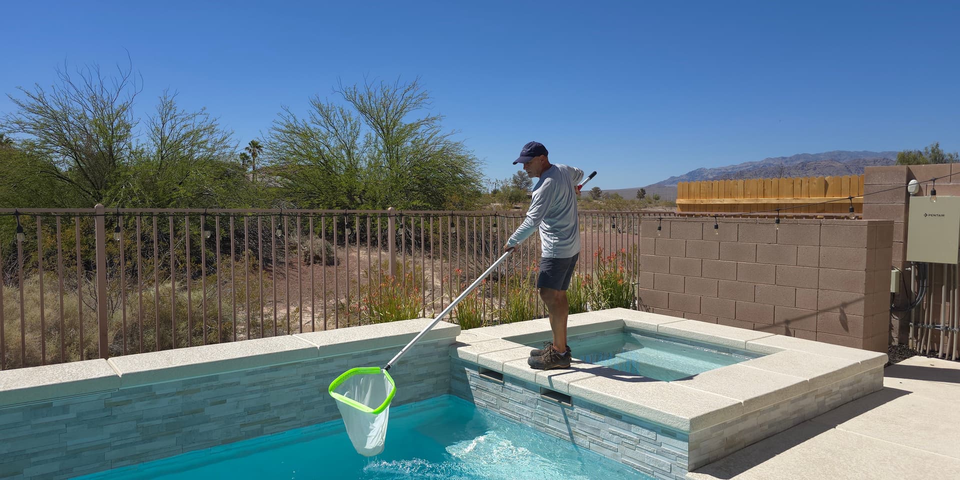 Michael Brown, AquaVision Pool Care Certified Pool Operator, skimming a residential pool and spa with desert vegetation and mountain backdrop
